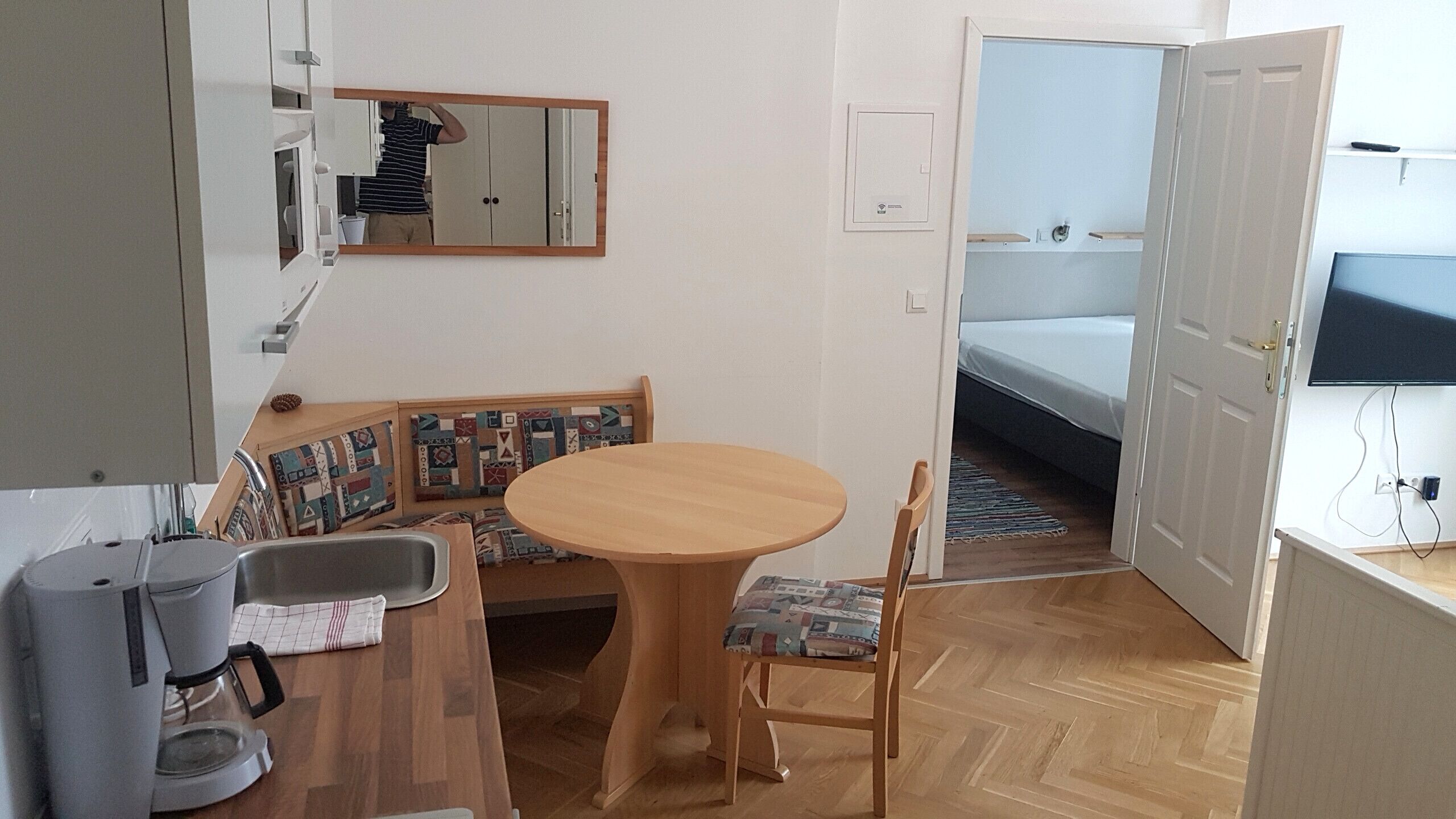A compact room with a wooden dining table, a bench with patterned cushions, a small kitchenette with a coffee maker, and a bed with a TV in the adjacent room.