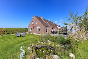Ein Backsteinhaus mit geneigtem Dach, ein Garten mit Teich und zwei Liegestühlen.