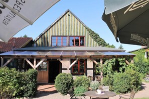 A building with a sloped roof, large windows, and a covered patio area with outdoor seating. - Holiday room 1 - RUMPELSTILZ baking and shower barn (Steinhagen)