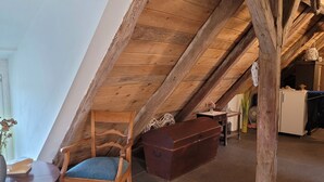 Un acogedor dormitorio en el ático, con techo de madera, una silla de madera con cojín, una mesita con un libro y un cofre marrón.