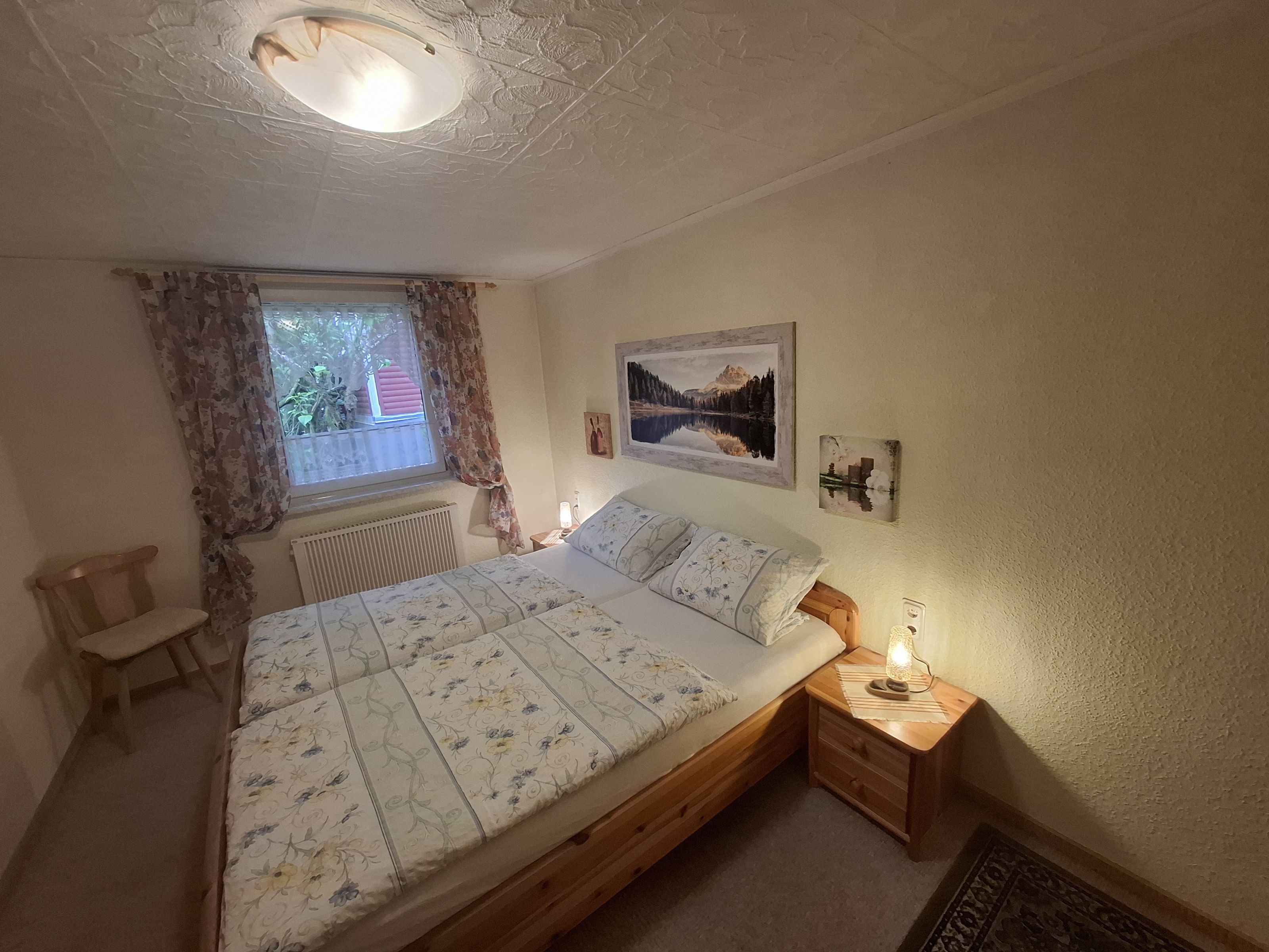 A bedroom with a wooden bed, a nightstand with a lamp, a window with curtains, and a radiator.