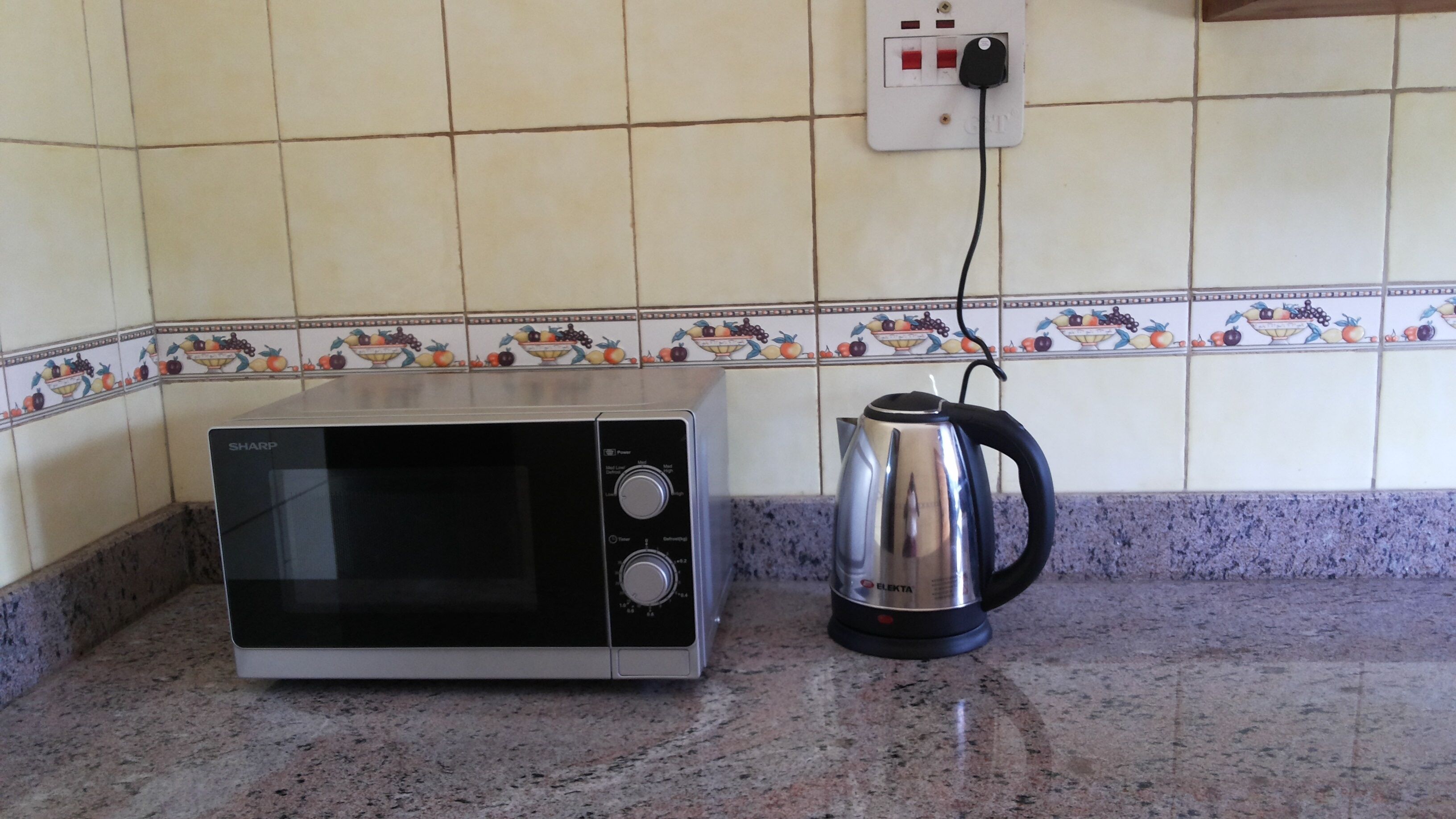 A kitchen with a microwave and a kettle on a granite countertop, a tiled backsplash with fruit designs, and a power outlet above.