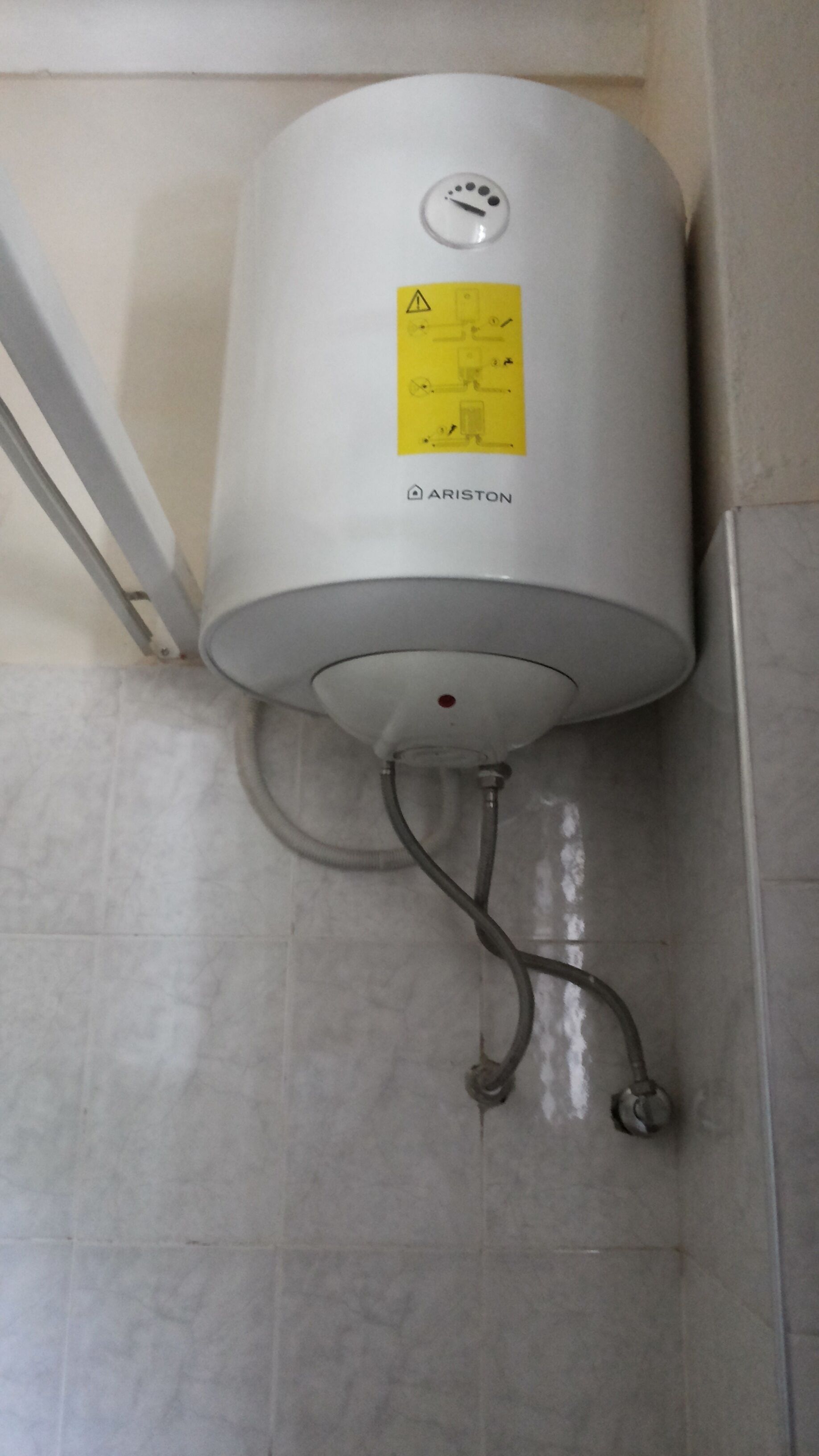 Ein weißer Boiler an einer gefliesten Wand, versehen mit einem gelben Warnhinweis.