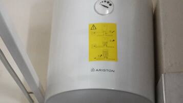 Ein weißer Boiler an einer gefliesten Wand, versehen mit einem gelben Warnhinweis.