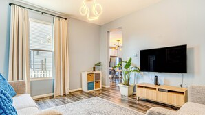 A modern living room with a flat-screen TV, a wooden cabinet, a potted plant, and a large window with curtains. - 1 Mi to Dtown & Lucas Oil Stadium: Fenced Yard (Indianapolis)