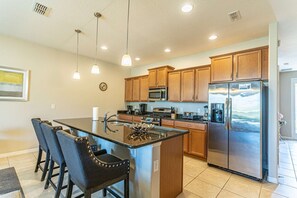 A modern kitchen with a central island, stainless steel appliances, wooden cabinets, pendant lighting, and a clock on the wall.
