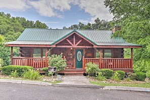 Eine rustikale Holzhütte mit einem grünen Metalldach, einer Veranda mit roten Geländern und einem kleinen Vorgarten mit Sträuchern und einem Briefkasten.