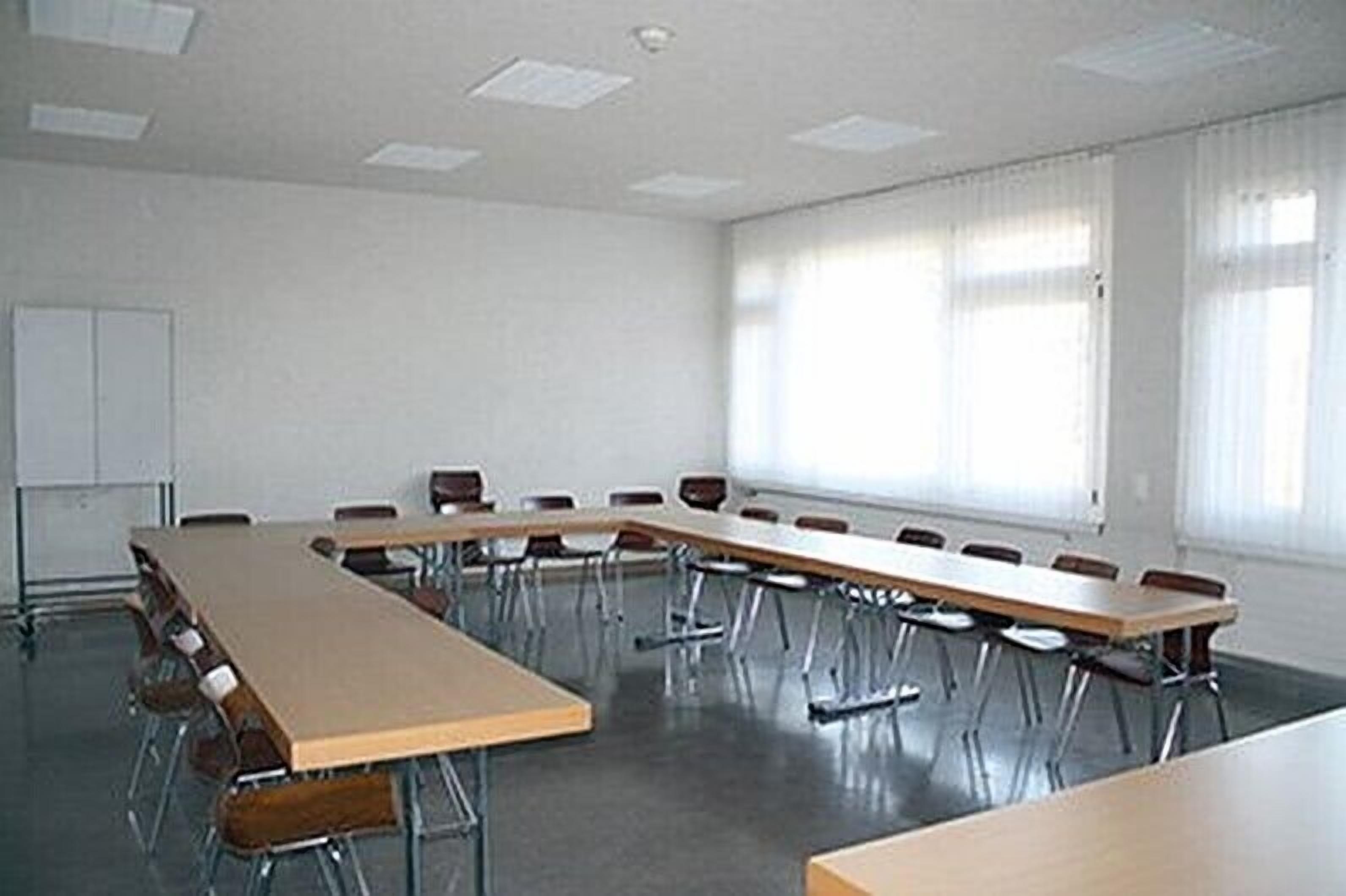 Un salón de conferencias con una larga mesa y sillas, ventanas amplias con persianas, y una pizarra en la pared.