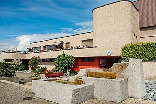 A modern building with a flat roof and a red section. There are potted plants and a small water feature in the foreground.