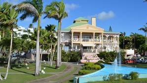 A large, two-story building with a green roof, surrounded by palm trees and a swimming pool with a fountain.