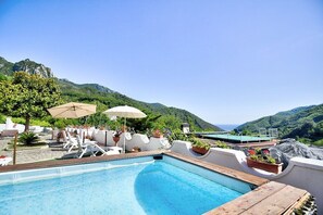 A rooftop pool with lounge chairs and umbrellas, surrounded by greenery and mountains.