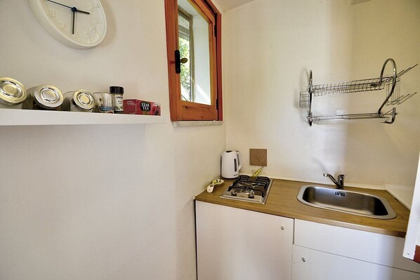 A small kitchen with a sink, stove, and a window. There are shelves with jars and a clock on the wall.