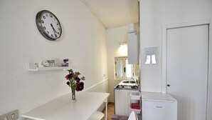 Ett litet pentry med diskho, spis och kylskåp. På väggen hänger en klocka och på bänkskivan står en vas med blommor.