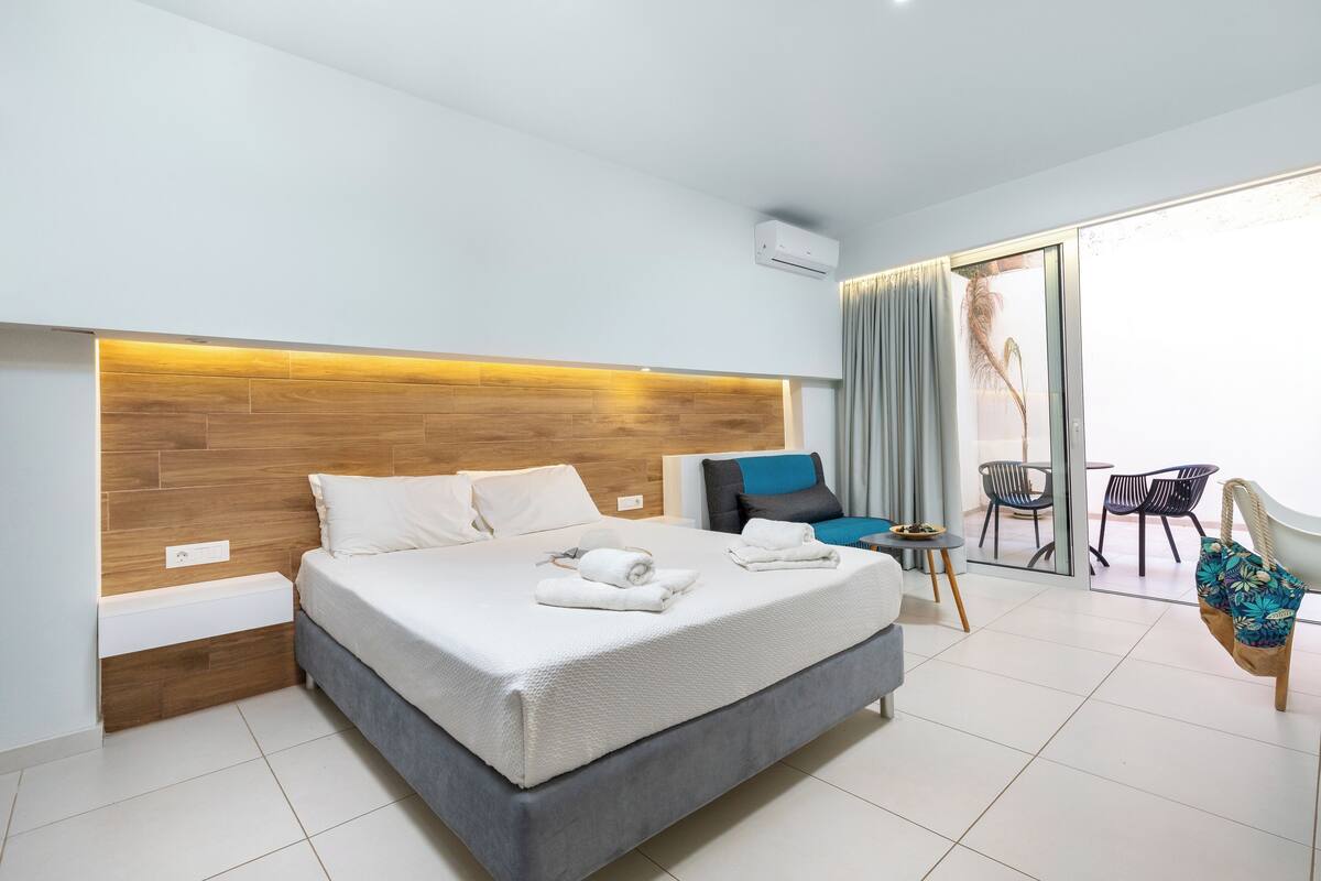 A modern hotel room with a large bed, a wooden headboard, a small bedside table, and a view of the outdoor area through a sliding glass door.