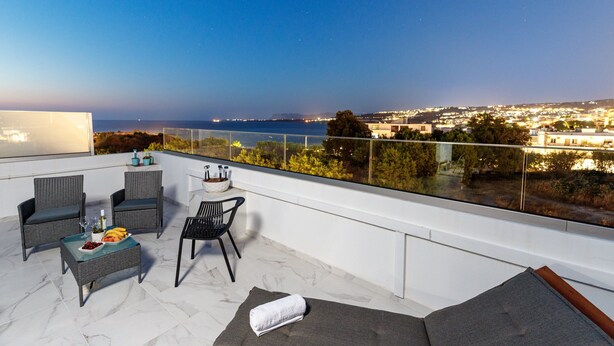 A balcony with wicker furniture, a glass table with food, and a view of the city and sea at night.
