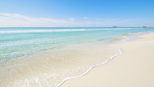 A beach with clear turquoise water, a sandy shore, and a pier extending into the distance.