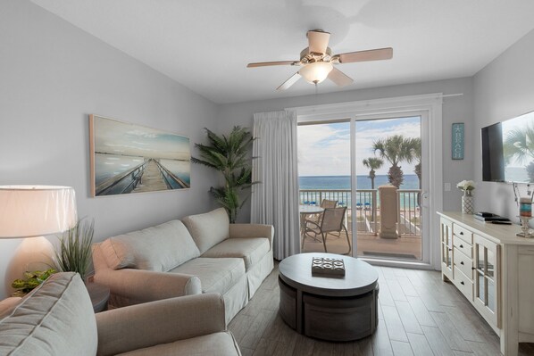 Un salón espacioso con un sofá grande, una mesa de centro y un ventilador de techo. Se aprecia una ventana amplia con vistas a la playa y palmeras.