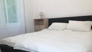 A neatly made bed with white bedding and a dark headboard in a room with a window and a nightstand with a lamp.