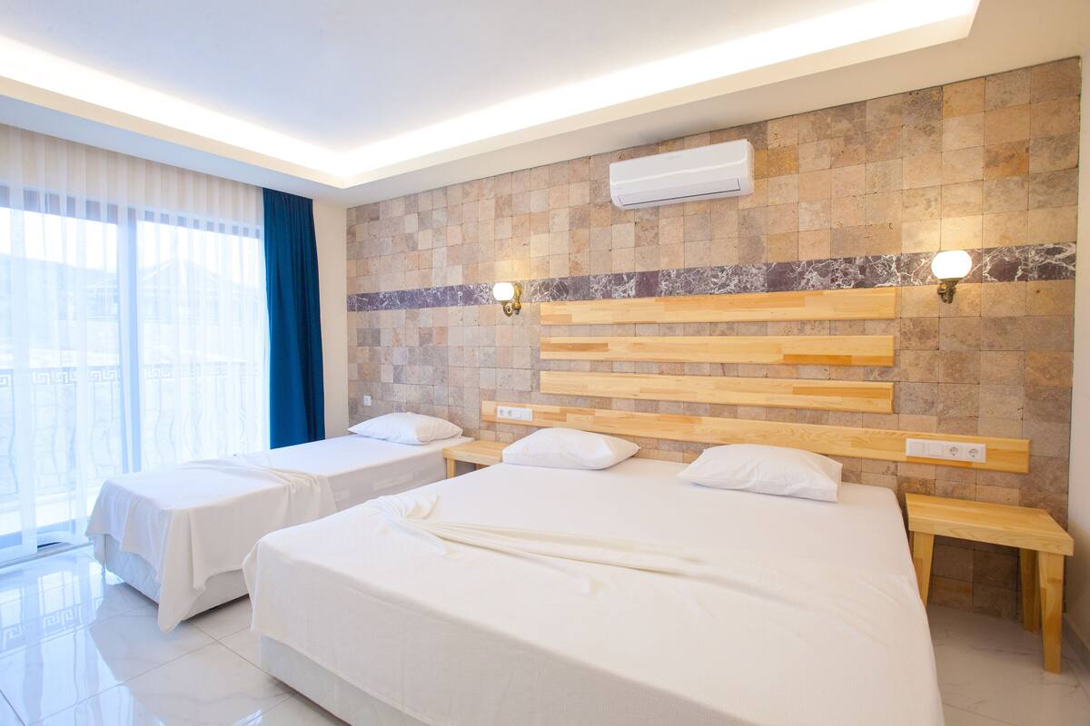 A hotel room with two beds, a large window with curtains, a marble-tiled wall, and an air conditioning unit.