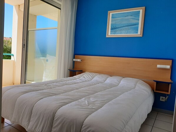 Un dormitorio con cama, ventana con cortinas, pared azul y un cuadro enmarcado.