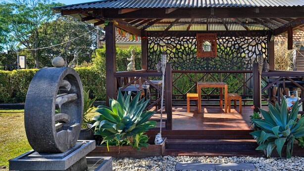Un kiosque en bois surmonté d’un toit en tôle ondulée, une sculpture en pierre, ainsi qu’une table et un banc en bois.