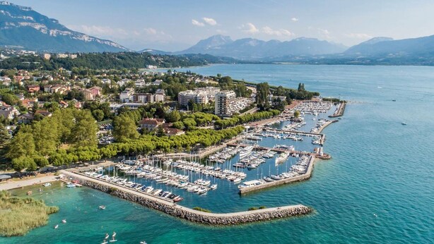 An aerial view of a coastal town with a marina, residential buildings, and mountains in the background.