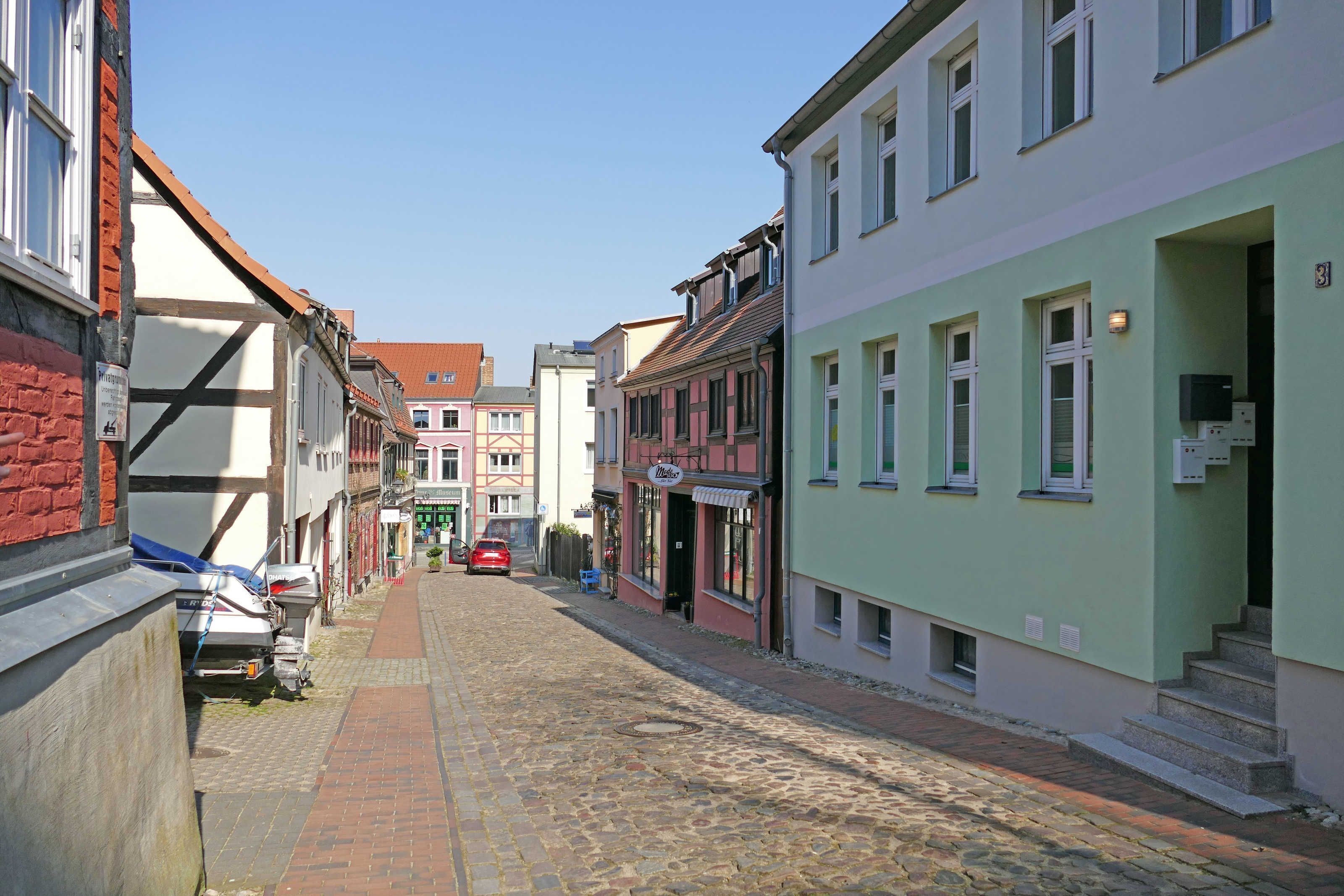 Una calle adoquinada flanqueada por edificios tradicionales europeos, un coche estacionado y un escaparate visible.