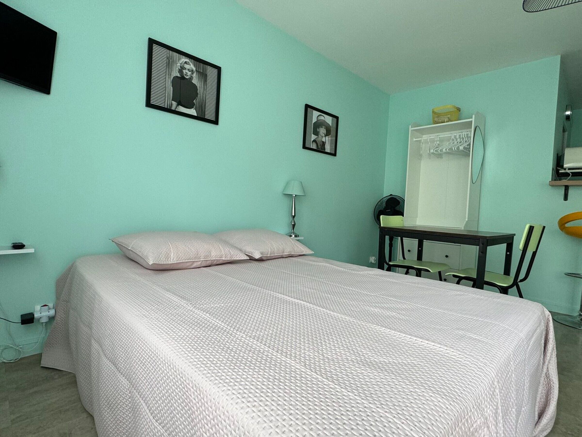 A bedroom with a bed, two framed pictures on the wall, a desk with a chair, and a mirror.
