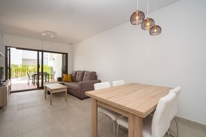 A modern living room with a wooden dining table, white chairs, a brown sofa, and a glass door leading to an outdoor area.