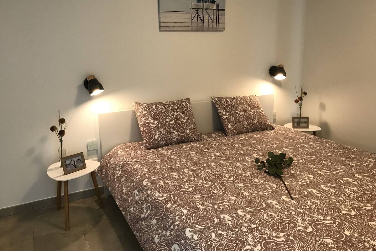 Ein Schlafzimmer mit einem großen Bett, zwei Nachttischen mit Lampen, einem gerahmten Bild auf einem Tisch und einem kleinen runden Tisch mit Vase und Bilderrahmen.