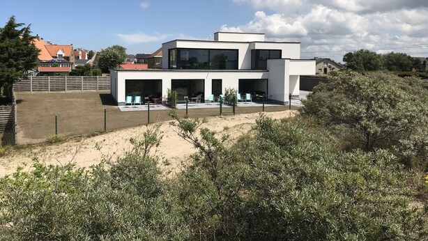 A modern two-story house with a flat roof, large windows, and a fenced outdoor area with chairs and tables. - In De Duinen (Middelkerke)