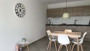 A modern kitchen with wooden cabinets, a round clock, a dining table with chairs, and a pendant light. - In De Duinen (Middelkerke)