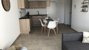 A modern kitchen with wooden cabinets, a dining area with white chairs, and a grey sofa. - In De Duinen (Middelkerke)