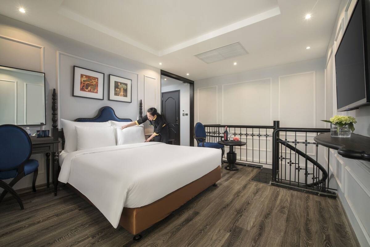 A modern hotel room with a large bed, a desk, a chair, a small table with a vase, and a balcony with a railing.