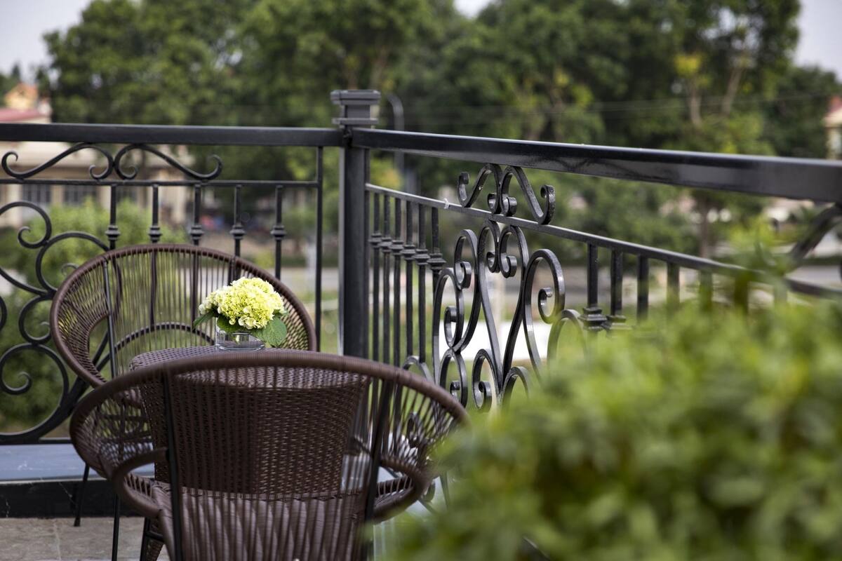 A balcony with wicker chairs and a table, a metal railing, and greenery in the background.