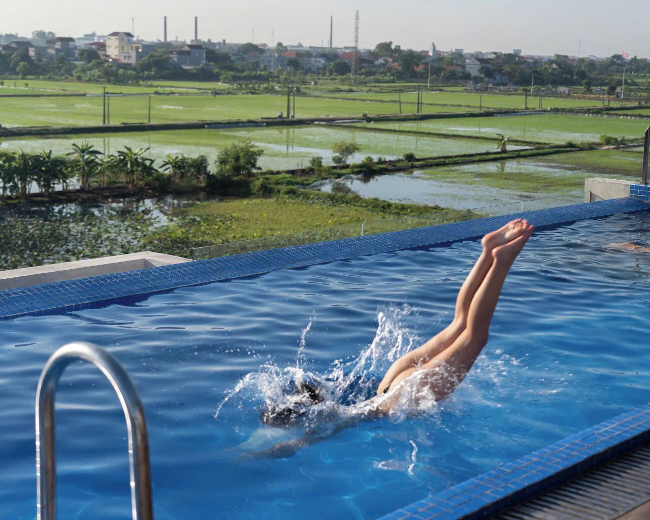 A swimming pool with a person diving into the water, surrounded by a view of green fields and distant buildings.