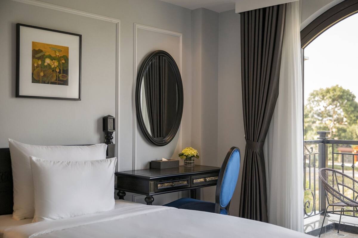 A hotel room with a bed, a desk with a vase of flowers, a large mirror, and a view of the outdoors.