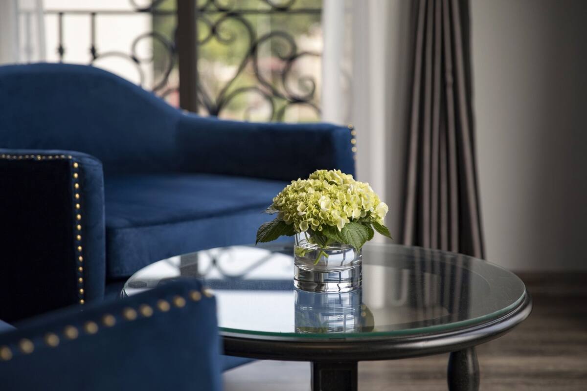 A room with a blue sofa, a glass coffee table with a vase of flowers, and a view of a balcony with a wrought iron railing.