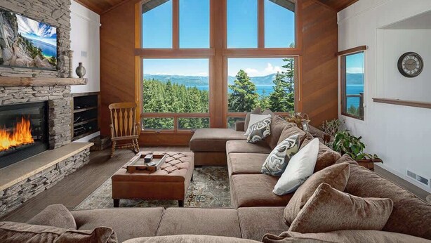 A cozy living room with a stone fireplace, a large window offering a view of trees and mountains, and a comfortable seating area. - Dynamic Views, Hot Tub, Gym, 2nd Living Room, Garage and More. (Carnelian Bay)