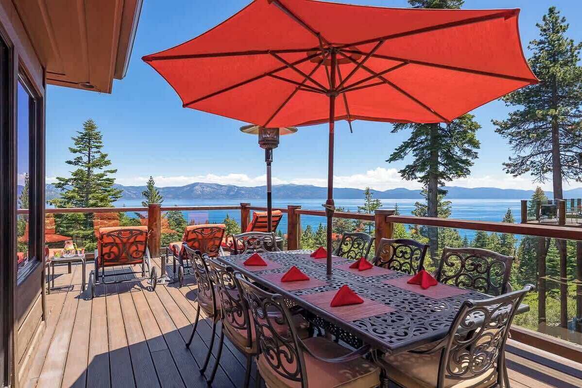 A wooden deck with a table and chairs, a red umbrella, and a view of mountains and a lake.