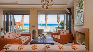 A spacious living room with a sofa set, a coffee table, and a fireplace. Large windows offer a view of the ocean and beach.