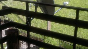 A balcony with a satellite dish, a trash bin, and a view of a grassy area and a fence.