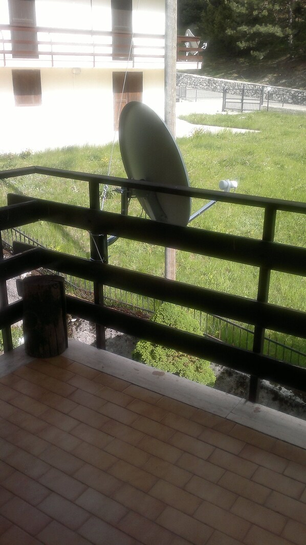 Un balcón con una antena parabólica, un bote de basura y vista a un área verde y una cerca.