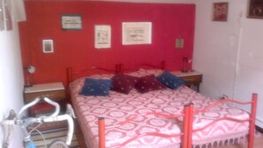 Un dormitorio con pared roja, una colcha estampada y varias almohadas. Se observan cuadros enmarcados en la pared y una bicicleta en la esquina.
