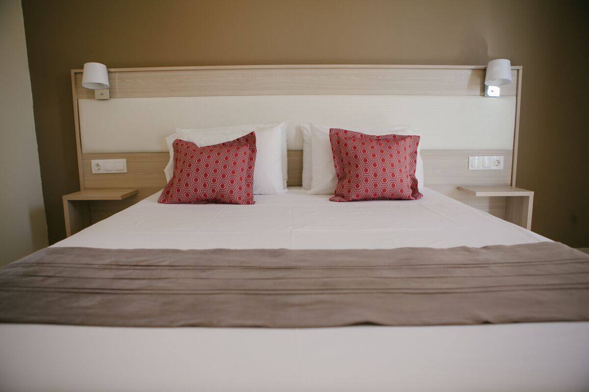 A neatly made bed with a white and brown bedspread, two patterned pillows, and two plain pillows. There are two bedside tables with lamps and a wall-mounted light fixture above the bed.