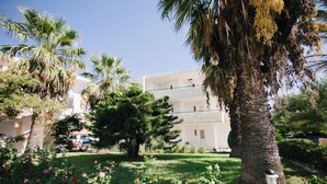 A building with a white facade, surrounded by lush greenery and palm trees. - Artemis Comfort & Pleasure (Rhodes)