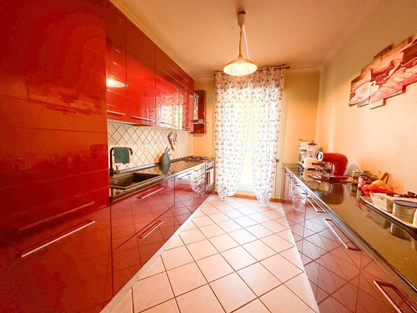 A kitchen with red cabinets, a tiled floor, a hanging light fixture, and a window with floral curtains.