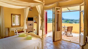 A bedroom with a four-poster bed, a desk, and a chair. There is a balcony with a table and a chair.