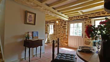 Un interior rústico con pared de piedra, vigas de madera, una silla de madera, una mesa con un jarrón de flores y una ventana con vistas a la vegetación.