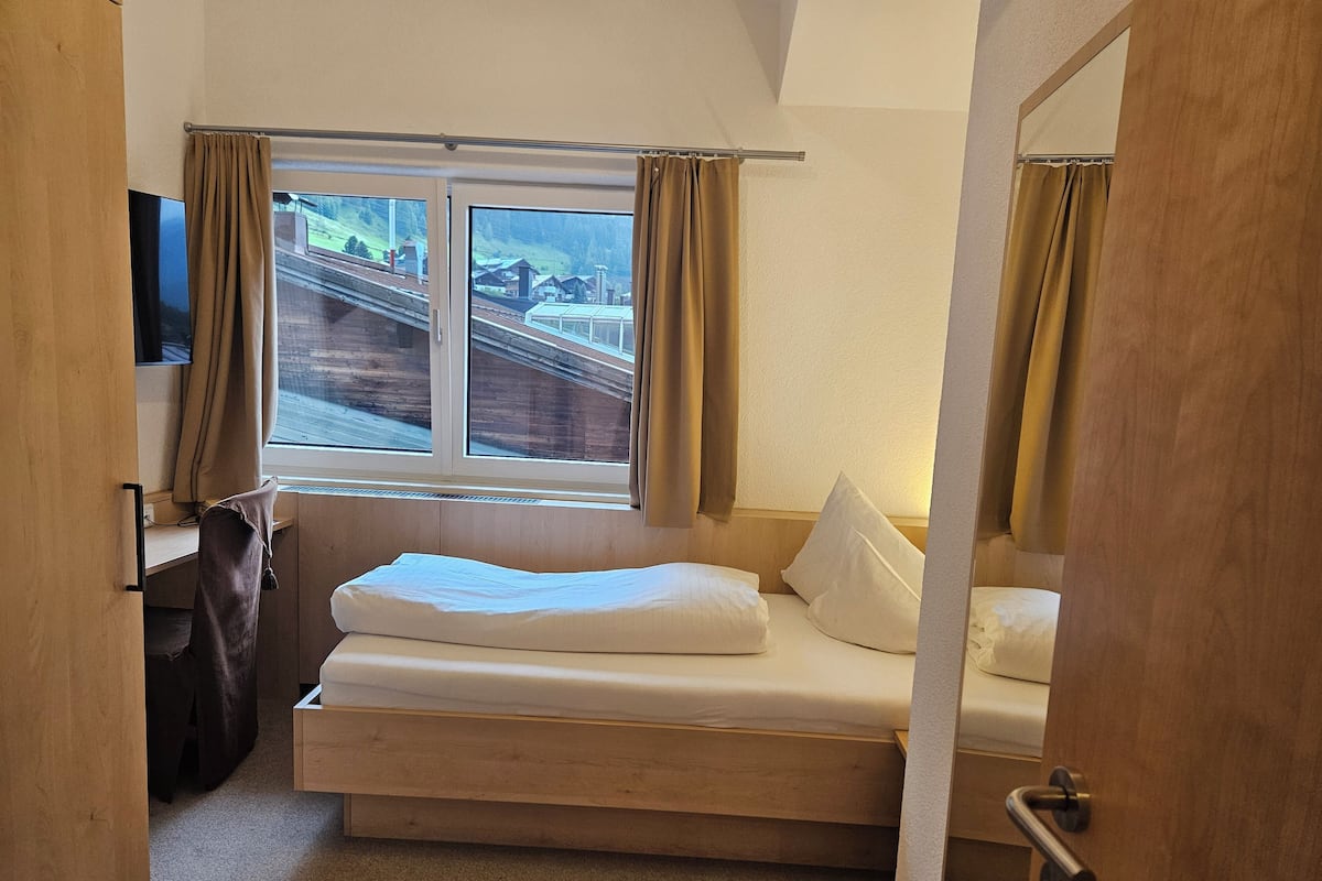 A small, compact room with a single bed, a window with curtains, a flat-screen TV, and a wooden door.